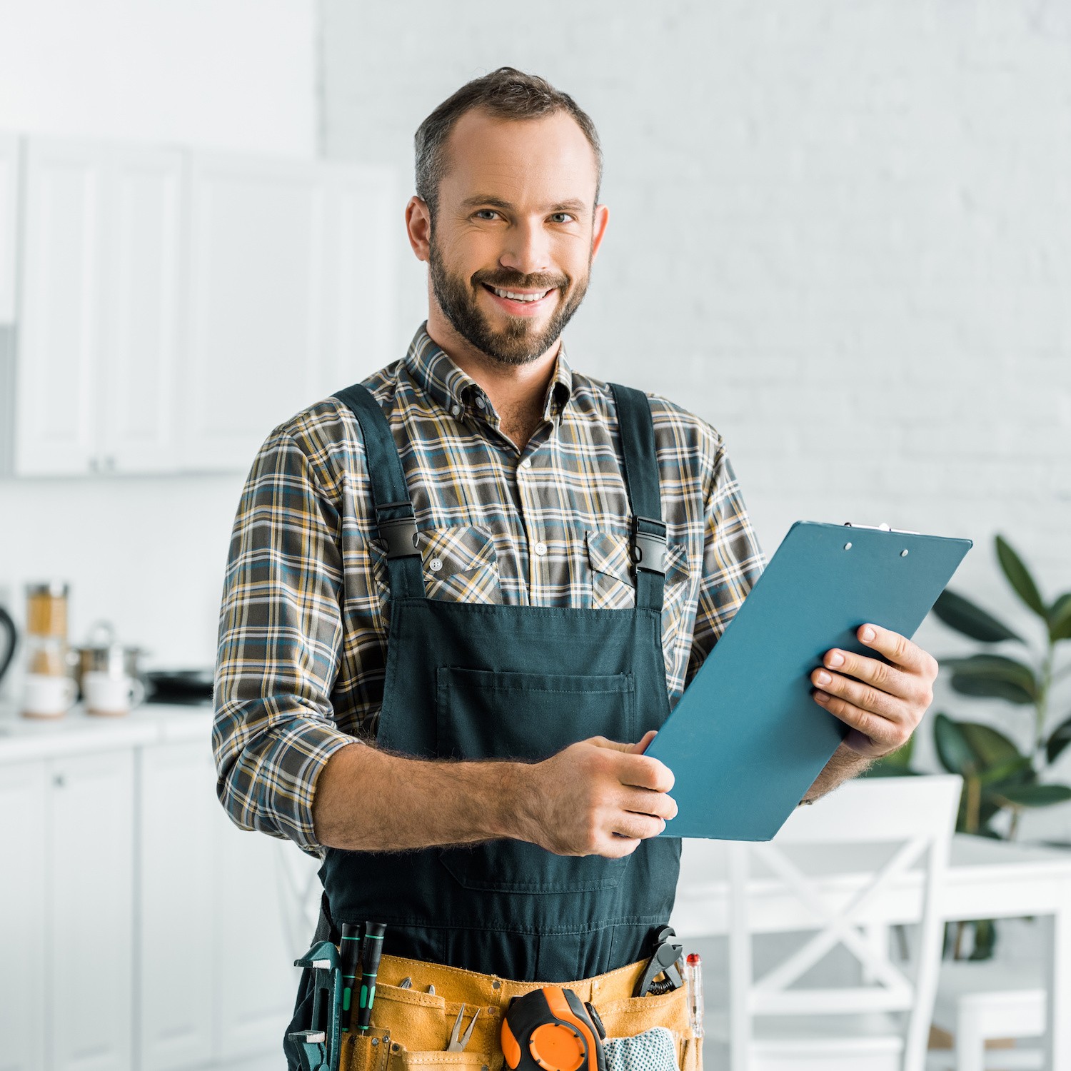 smiling handsome plumber holding clipboard and looking at camera in kitchen